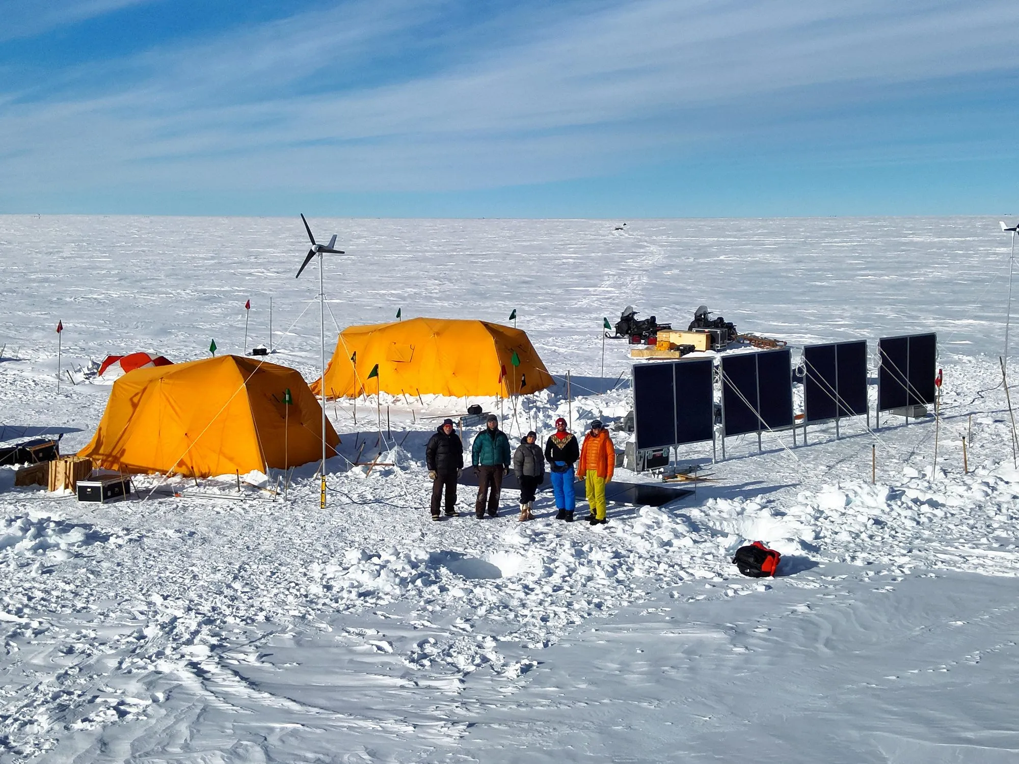 A photo of the install team in front of the camp tents, solar panels, and wind turbines, under blue sky